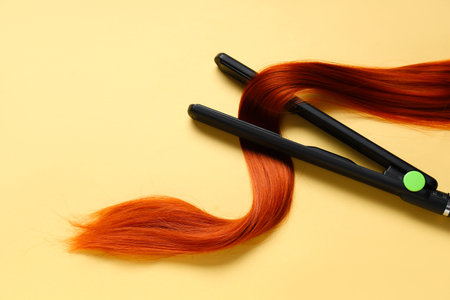 Hair straightener with red hair strand on pale yellow background