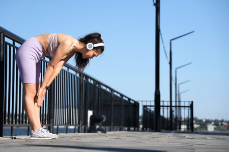 Beautiful young tired sporty Asian woman in headphones resting while jogging along waterfront...