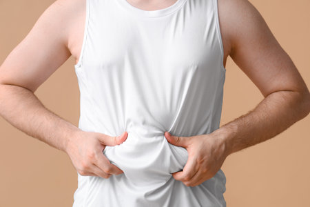 Young overweight man touching fat on his belly against beige background, closeup. Weight gain...