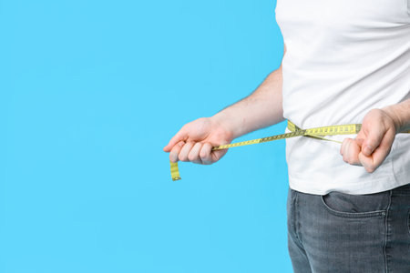 Young overweight man with measuring tape on blue background, closeup. Weight gain conceptの写真素材