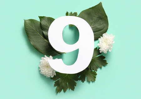 Green leaves and white flowers with paper number nine on turquoise background. Top view
