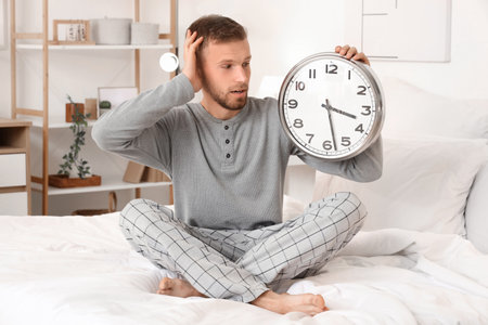 Young man holding clock and sitting on bed at home. Insomnia conceptの写真素材