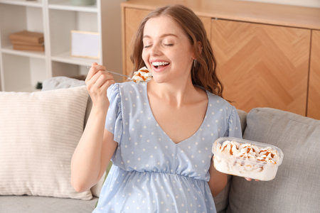 Young pregnant woman eating ice cream on sofa at homeの写真素材