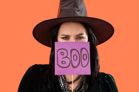 Beautiful young woman dressed as witch for Halloween with the word BOO on orange backgroundの写真素材