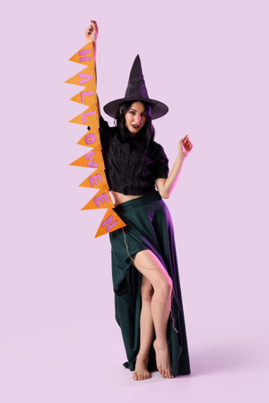 Beautiful young happy woman dressed as witch for Halloween with garland on lilac backgroundの写真素材