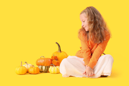 Cute little happy girl in autumn clothes with different pumpkins sitting on yellow backgroundの写真素材
