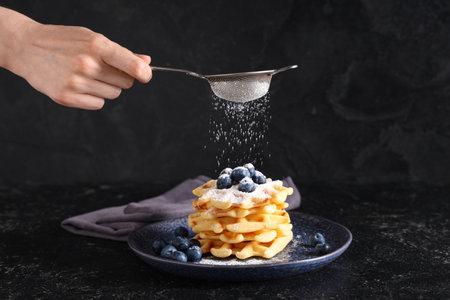 Woman sprinkling tasty homemade waffles with sugar powder on black backgroundの写真素材