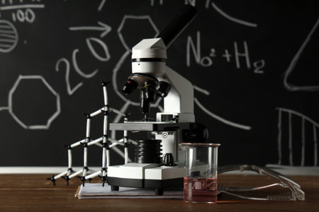 Modern microscope, molecular model and flask on wooden table near chalkboard with chemical formulas
