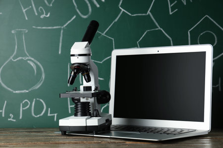 Modern microscope and laptop on wooden table near chalkboard with chemical formulasの写真素材