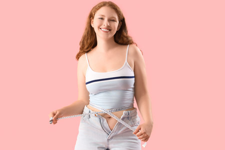 Young woman in tight shorts measuring her waist on pink background. Weight gain concept