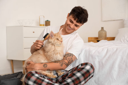 Handsome young happy man brushing cute Maine Coon cat in bedroom at homeの写真素材