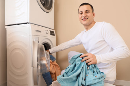 Young man putting dirty clothes into washing machine in laundry room