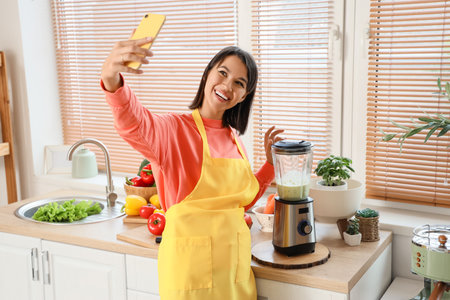 Young woman with mobile phone and blender taking selfie in kitchenの写真素材