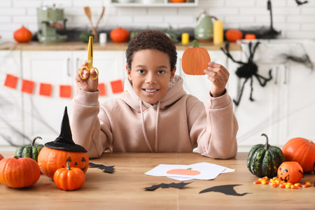 Teenage African-American boy with paper Halloween pumpkin and scissors in kitchenの写真素材