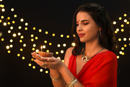 Beautiful young Indian woman in traditional sari with diya lamp against blurred lights on black background. Divaly celebrationの写真素材