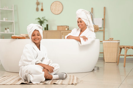 Happy young African-American mother and her daughter in bathrobes sitting inside bathtub at home