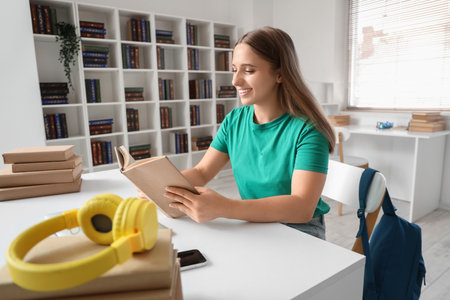Happy female student with headphones reading book while studying at libraryの写真素材