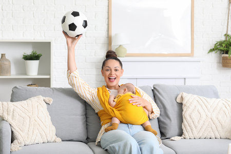 Young mother with little baby in sling and soccer ball watching football at homeの写真素材