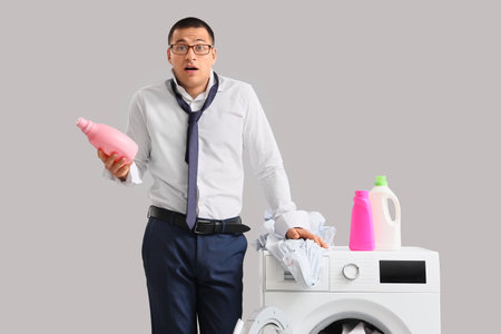 Confused young businessman with bottles of laundry detergent on washing machine against gray backgroundの写真素材