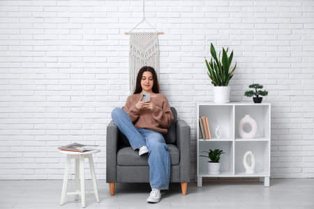 Young woman using mobile phone on black armchair in living roomの写真素材