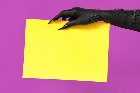 Black hand of witch with yellow paper sheet on purple background. Halloween celebrationの写真素材