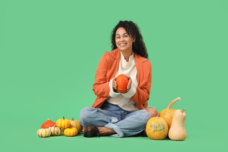 Beautiful young happy African-American woman with pumpkins sitting on green backgroundの写真素材