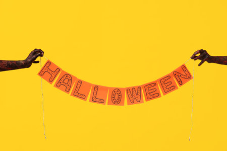 Hands of zombies holding paper garland with the word HALLOWEEN on yellow backgroundの写真素材