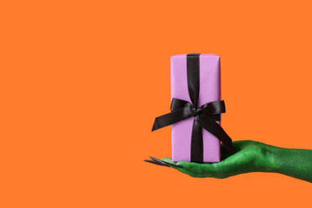 Green hand of witch with gift box on orange background. Halloween celebrationの写真素材