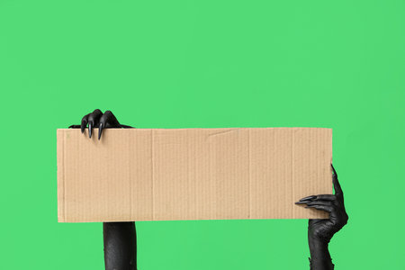 Black hands of witch with blank cardboard sheet on green background. Halloween celebrationの写真素材