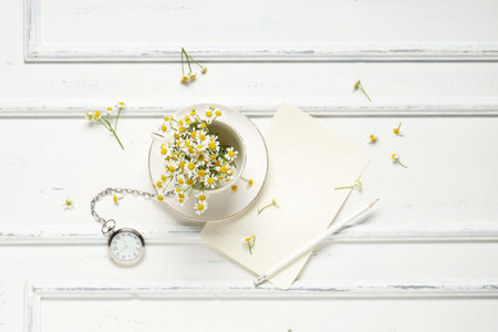 Composition with ceramic cup, chamomile flowers, blank paper sheet and pocket watch on light backgroundの写真素材