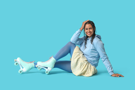 Beautiful young African-American woman in roller skates with dreadlocks sitting on blue backgroundの写真素材