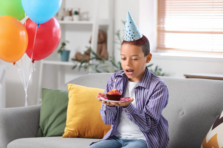 Cute little African-American boy in party hat with birthday cake sitting on sofa in living room