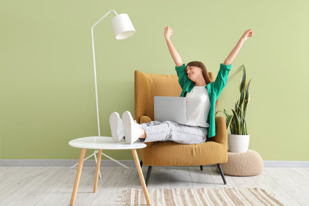 Happy young woman with laptop sitting in armchair near green wallの写真素材