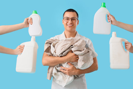 Young man holding dirty clothes and hands with bottles of laundry detergent on blue backgroundの写真素材