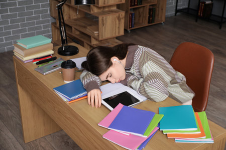 Tired female student with earphones and tablet computer sleeping at table in libraryの写真素材