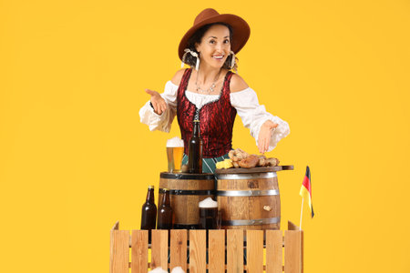 Beautiful mature happy Octoberfest waitress with barrels, bottles of beer and snacks on table against yellow backgroundの写真素材