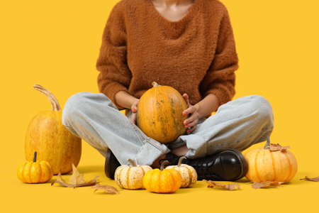 Beautiful young woman with pumpkins and autumn leaves sitting on yellow background, closeupの写真素材