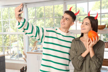 Young happy couple with pumpkin taking selfie in kitchen decorated for Halloween party