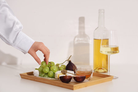 Female hand with sweet ripe grapes, figs, camembert cheese, honey and bottle of wine on white...