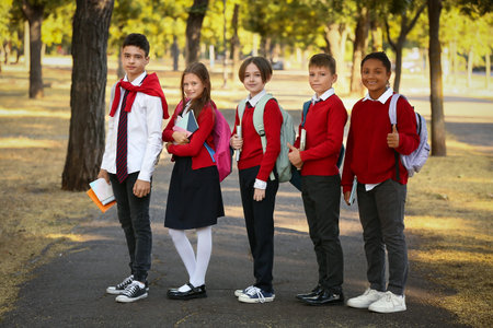 Group of happy pupils with stationery showing thumbs-up gesture while going to school outdoors
