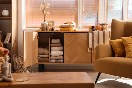 Open commode with stacks of stylish clothes and autumn decorations near window in living roomの写真素材
