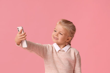 Cute little happy girl in warm stylish sweater with mobile phone taking selfie on pink background