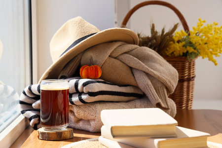 Stack of stylish autumn clothes with books and glass of coffee on table near window at homeの写真素材