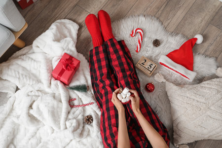 Young woman in pajamas with cup of cocoa, gift box and Christmas decorations on floor at home, top viewの写真素材