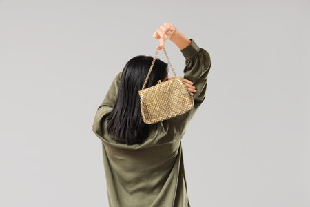 Beautiful young woman with stylish golden bag on gray background, back view