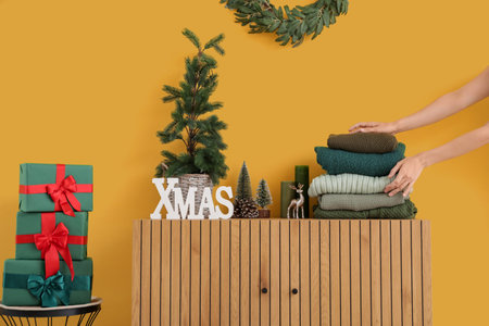 Woman putting stack of winter clothes on wooden cabinet with Christmas decorations near yellow wallの写真素材