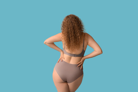 Beautiful young body positive woman in stylish underwear on blue background, back view