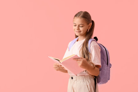 Cute schoolgirl reading book with backpack on pink backgroundの写真素材