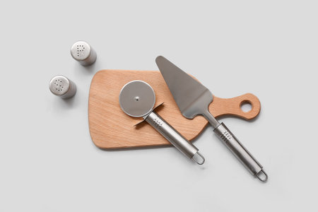 Wooden board with steel spatula, pizza cutter and spices on gray background