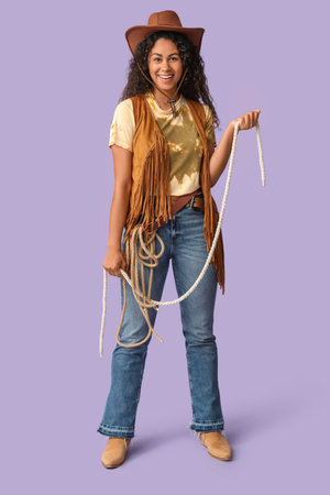 Beautiful African-American cowgirl with lasso on lilac backgroundの写真素材